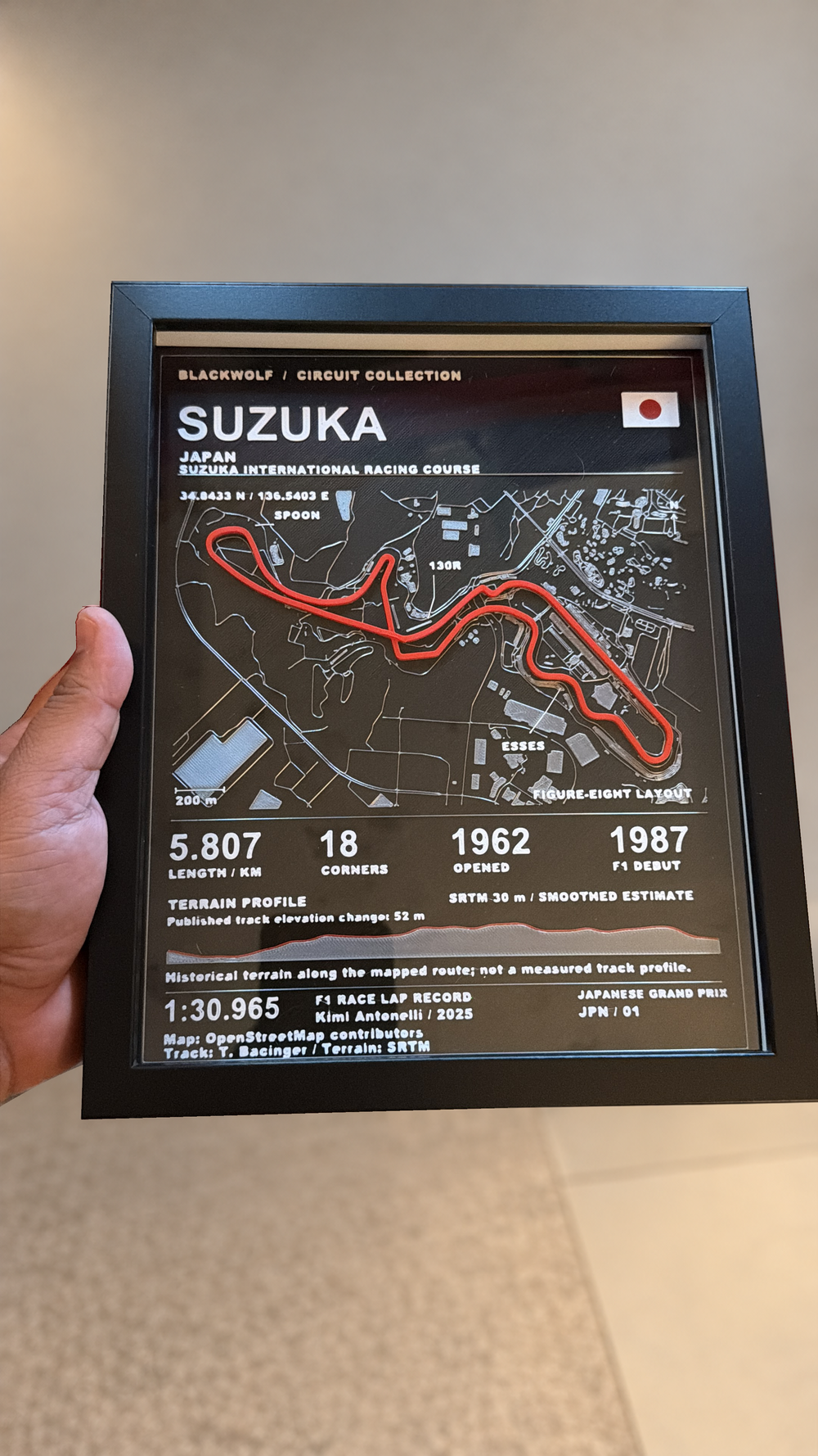 Photograph of the real Suzuka circuit artwork in a black frame held in hand; background cleaned, artwork and hand preserved.