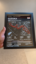 Photograph of the real Suzuka circuit artwork in a black frame held in hand; background cleaned, artwork and hand preserved.